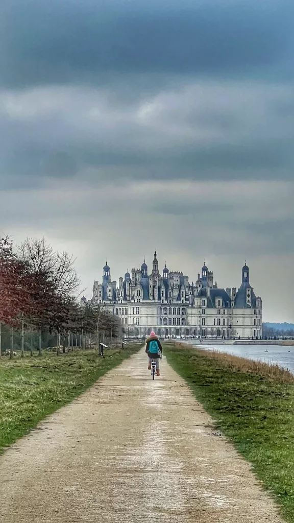 Vista del camino hacia el Château de Chambord en el Valle del Loira, Francia, con una persona en bicicleta y la imponente fachada renacentista al fondo.